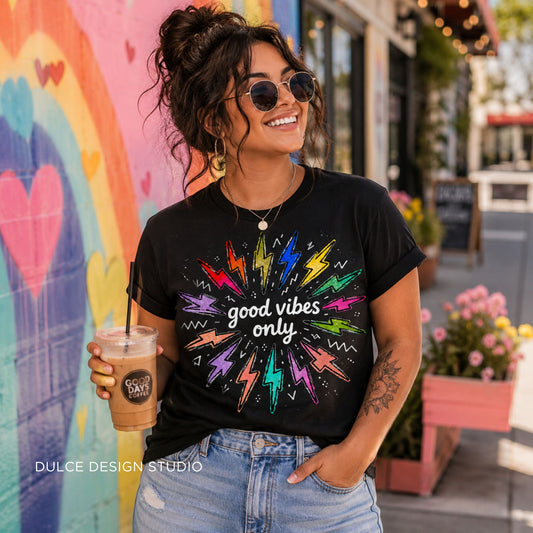 A woman wearing a black graphic t-shirt with a colorful lightning-bolt burst design and the text 'good vibes only', paired with light-wash jeans and holding an iced coffee in front of a colorful mural.