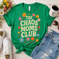 A bright green graphic T-shirt laid flat on a light wood surface, styled with jeans, white sneakers, a coffee mug, and flowers. The shirt reads “THE CHAOS MOMS CLUB” in large cream letters with colorful floral accents.