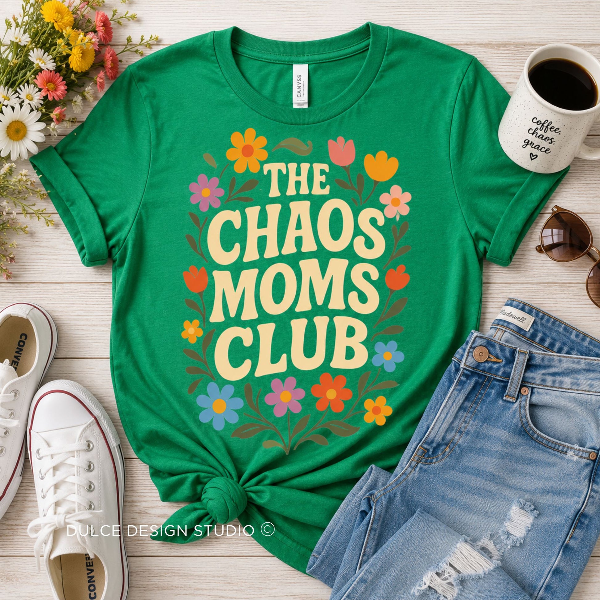 A bright green graphic T-shirt laid flat on a light wood surface, styled with jeans, white sneakers, a coffee mug, and flowers. The shirt reads “THE CHAOS MOMS CLUB” in large cream letters with colorful floral accents.