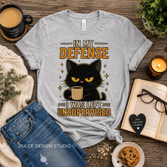 A heather gray graphic T-shirt laid flat on a rustic wooden surface, styled with a coffee mug, book, glasses, cookie, knit fabric, and a candle. The shirt features a black cat illustration holding a coffee cup with the text 'IN MY DEFENSE I WAS LEFT UNSUPERVISED.