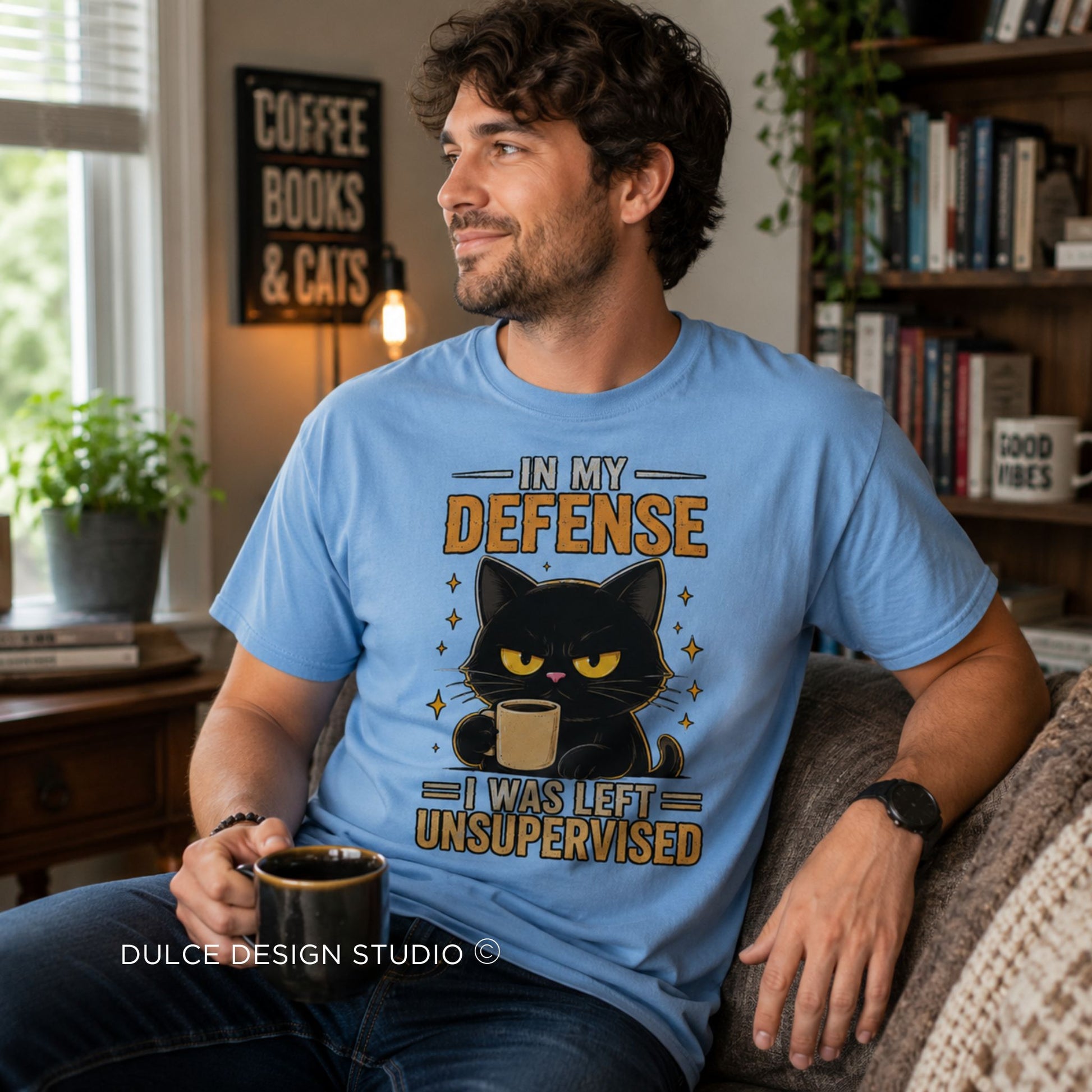 A man sitting on a couch in a cozy living room wearing a light blue graphic t-shirt. The shirt features a cartoon black cat with yellow eyes holding a mug, with the text 'IN MY DEFENSE I WAS LEFT UNSUPERVISED'.