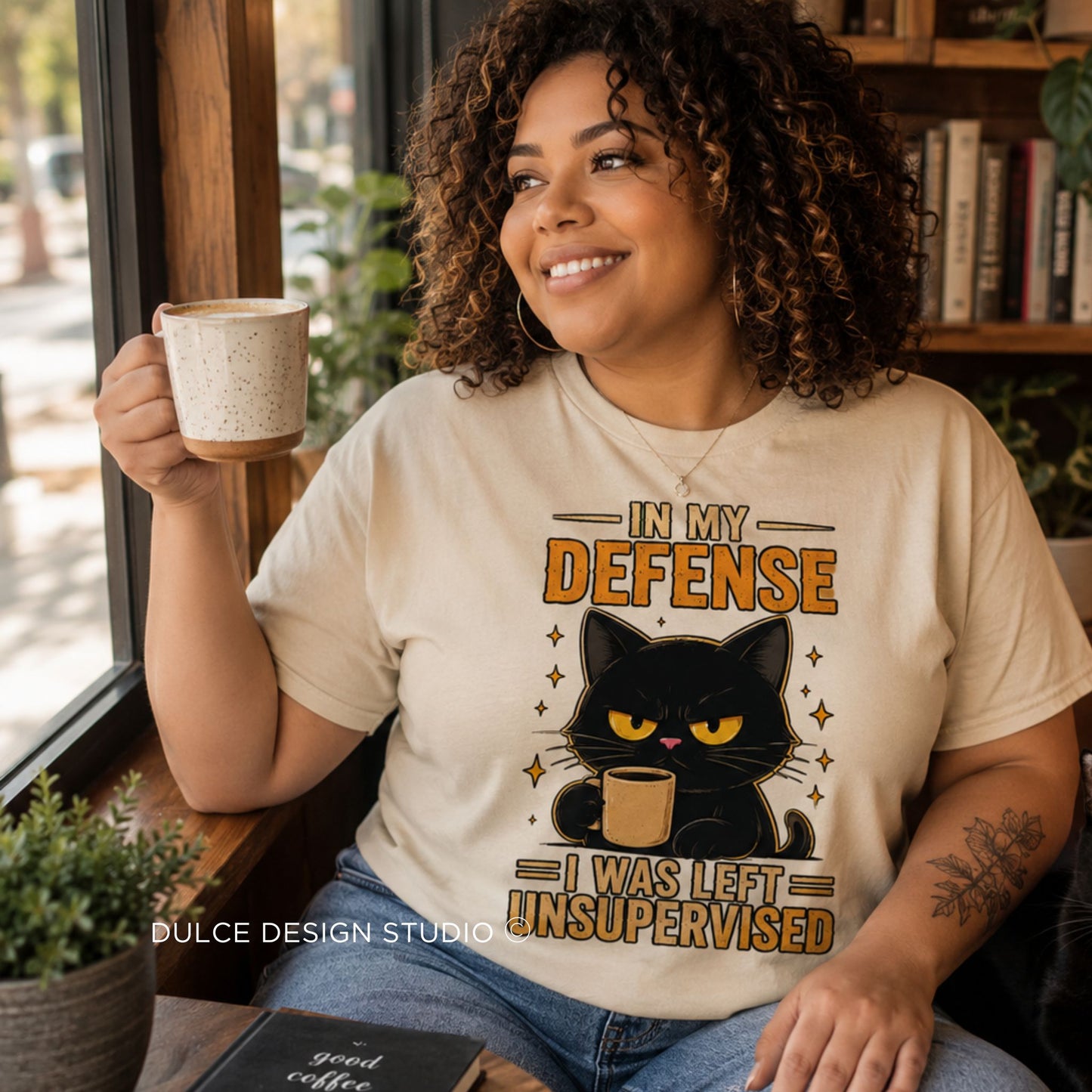 A woman sitting indoors by a window wears a cream graphic T-shirt with a black cat illustration and the text 'IN MY DEFENSE I WAS LEFT UNSUPERVISED.' She holds a speckled mug, with shelves, plants, and a book visible in the background.