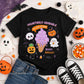 A black short-sleeve graphic T-shirt laid flat on a white wood surface with Halloween props around it. The shirt features a colorful cartoon design with a purple ice cream cone, jack-o'-lanterns, ghosts, candy corn, stars, and the text 'HAUNTINGLY ADORABLE' and 'BOOS! BREWS!'.