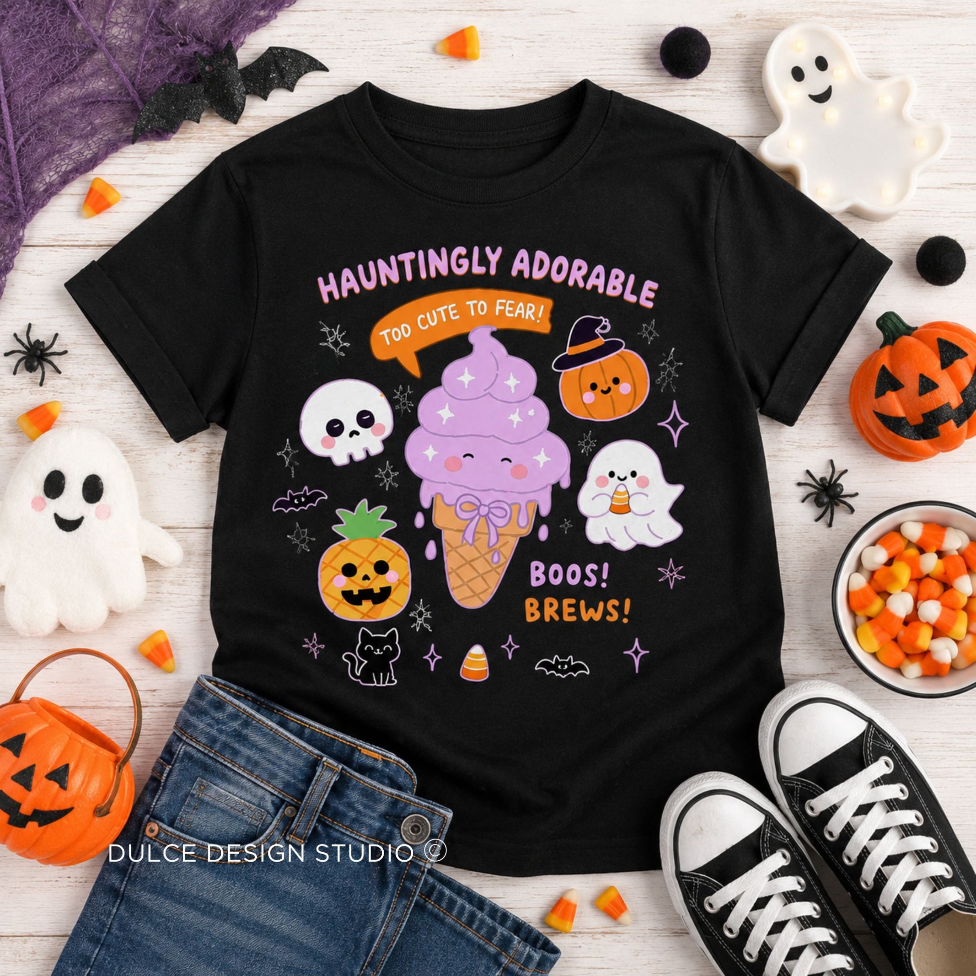 A black short-sleeve graphic T-shirt laid flat on a white wood surface with Halloween props around it. The shirt features a colorful cartoon design with a purple ice cream cone, jack-o'-lanterns, ghosts, candy corn, stars, and the text 'HAUNTINGLY ADORABLE' and 'BOOS! BREWS!'.