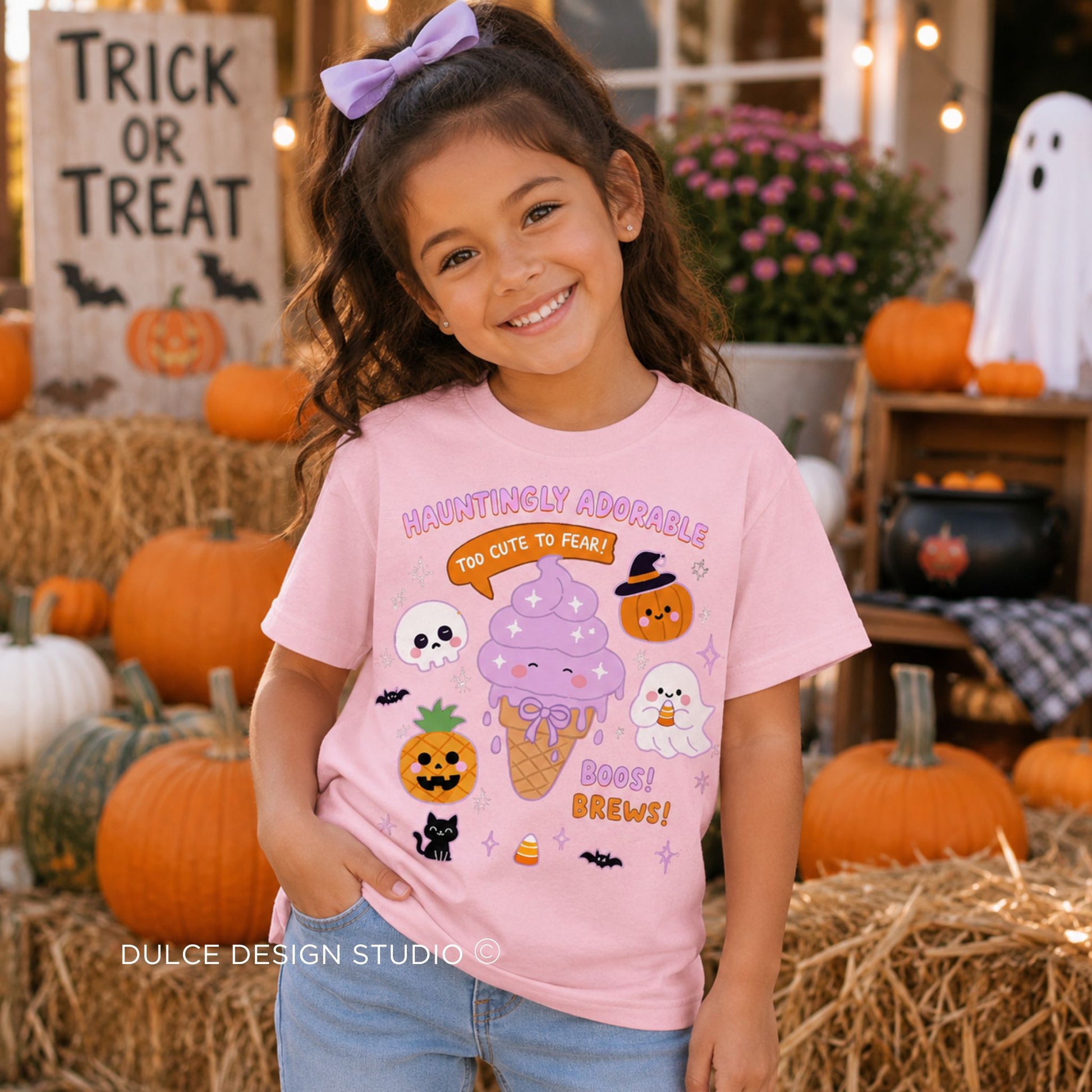 A young girl with a purple hair bow wearing a light pink graphic t-shirt with a cute Halloween theme, standing in a cozy autumn-themed setting with pumpkins and hay bales.
