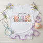White baby onesie with 'Heart of a Healer Nurse' text, surrounded by flowers and medical accessories on a light wooden background.