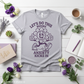 Gray t-shirt with a purple graphic and text on a white wooden surface with flowers and a cup of coffee.