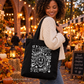 A woman wearing a cream knit sweater and jeans carries a black canvas tote bag with white Halloween-themed illustrations and text, set against a warm, autumnal outdoor market background.