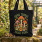 A black canvas tote bag with a colorful mushroom and floral graphic and the words “GOOD THINGS TAKE TIME,” photographed outdoors on a mossy stone in a garden setting. The tote bag is the product, and the background is a real outdoor environment.