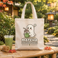 A beige canvas tote bag with a cute ghost graphic holding a green matcha drink, with the text 'MATCHA IS MY HAPPY PLACE' printed below. The bag is displayed on a bamboo table in an outdoor garden setting with a matcha iced coffee, a bamboo whisk, and autumn leaves.