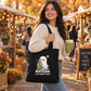 A woman in a cream knit sweater and jeans stands in an outdoor autumn market holding a green matcha drink and carrying a black tote bag with a cute ghost graphic and the text 'MATCHA IS MY HAPPY PLACE'.