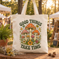 A natural canvas tote bag with a colorful mushroom and floral graphic and the text 'GOOD THINGS TAKE TIME' in dark green, displayed on a wooden table in an outdoor market setting.