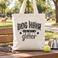 A natural canvas tote bag with the text 'DOG HAIR IS MY glitter' and paw prints, displayed on a wooden bench outdoors with a dog leash, tennis ball, and metal bowl nearby.