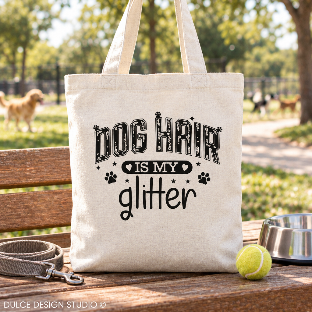 A natural canvas tote bag with the text 'DOG HAIR IS MY glitter' and paw prints, displayed on a wooden bench outdoors with a dog leash, tennis ball, and metal bowl nearby.