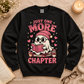 Black crewneck sweatshirt flat lay featuring a cute ghost reading a book with Just One More Chapter text.