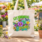 A natural canvas tote bag with a colorful 'LET KINDNESS BLOOM' graphic featuring flowers, butterflies, and hearts, displayed on a wooden table in a sunny flower garden.