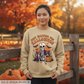 A woman stands outdoors in front of a wooden fence and pumpkins, wearing a beige crewneck sweatshirt with a Halloween graphic of a skeleton and the text 'JUST WAITING FOR HALLOWEEN'. The background features warm autumn foliage and pumpkins.