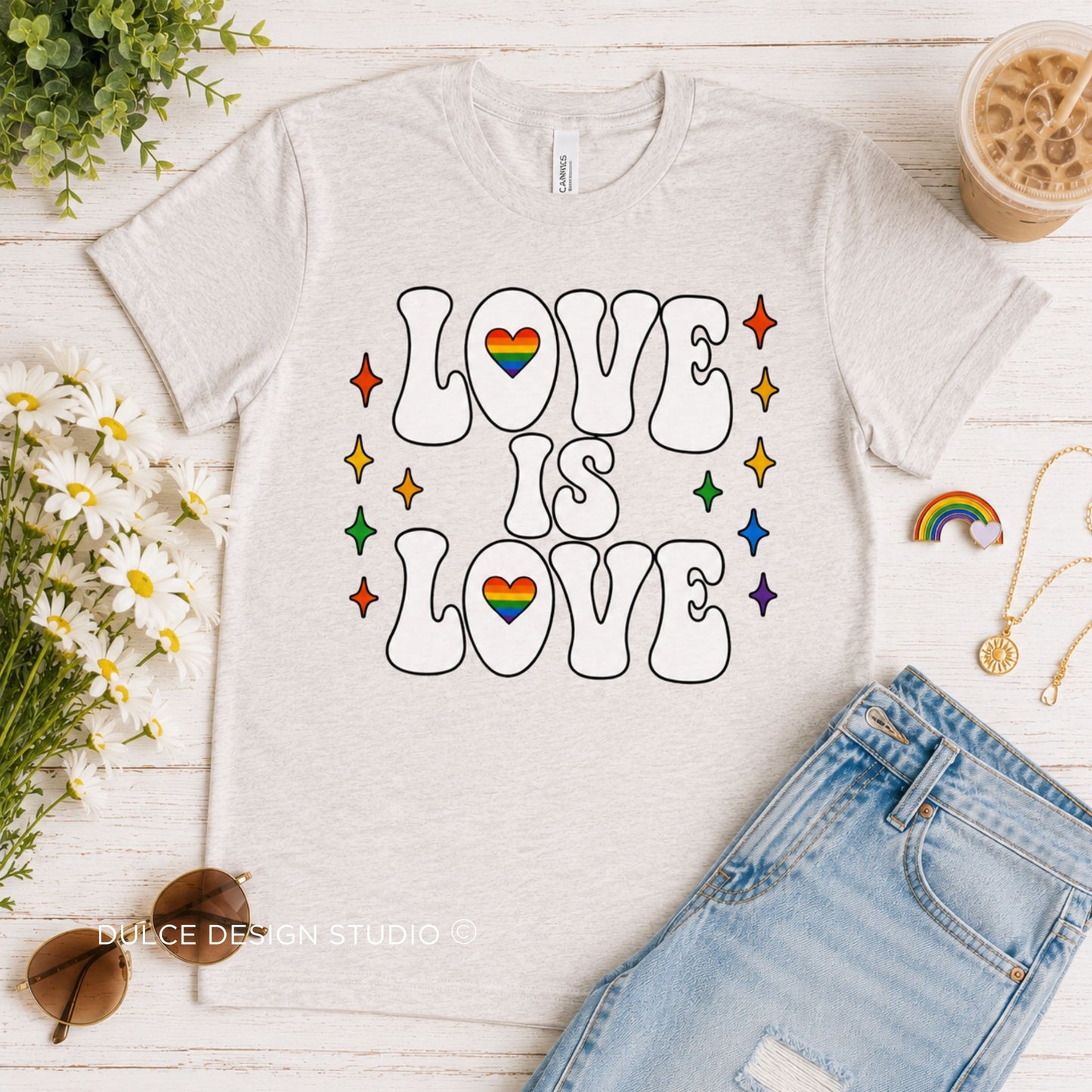White t-shirt with 'LOVE IS LOVE' text and rainbow hearts on a wooden surface with flowers and sunglasses.