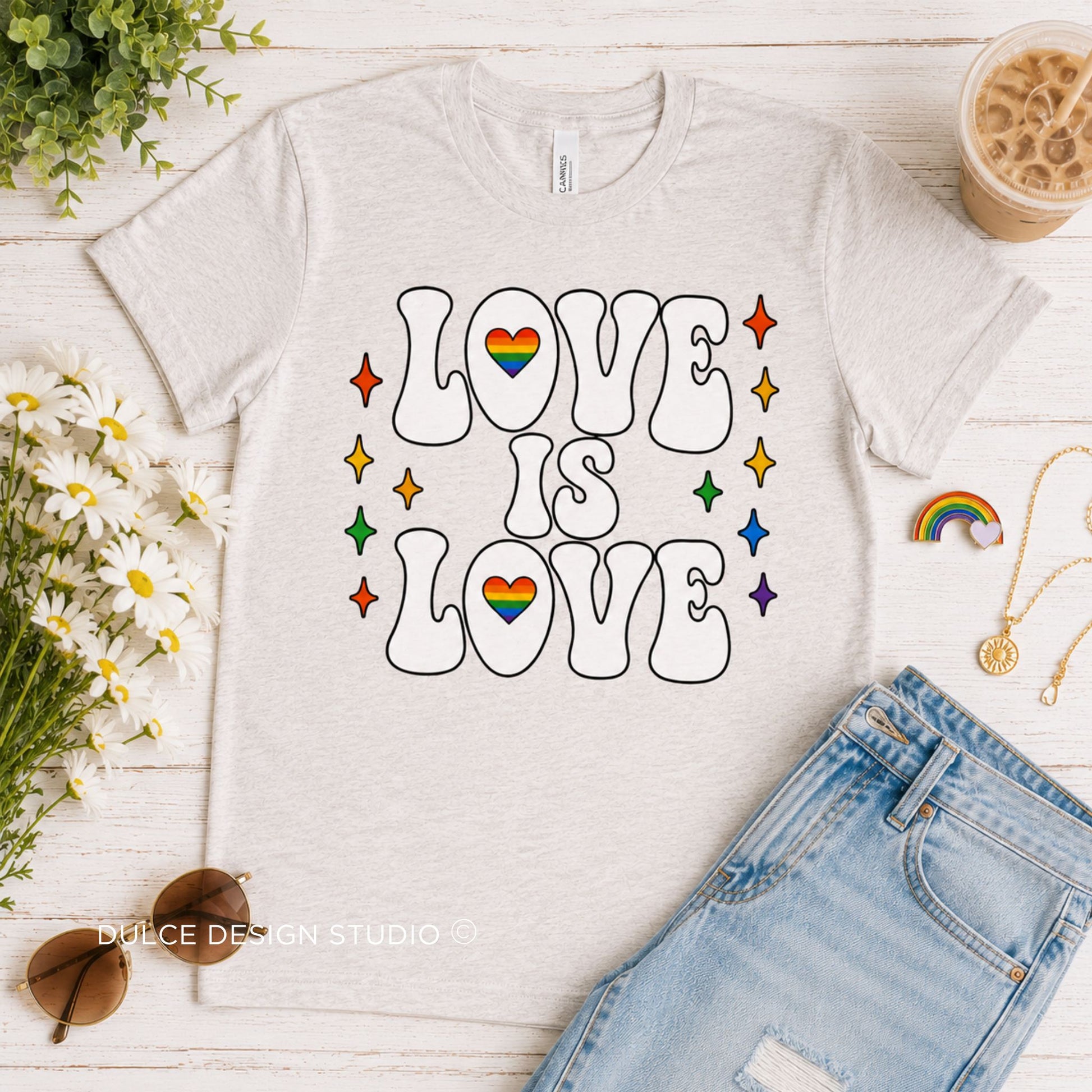 White t-shirt with 'LOVE IS LOVE' text and rainbow hearts on a wooden surface with flowers and sunglasses.