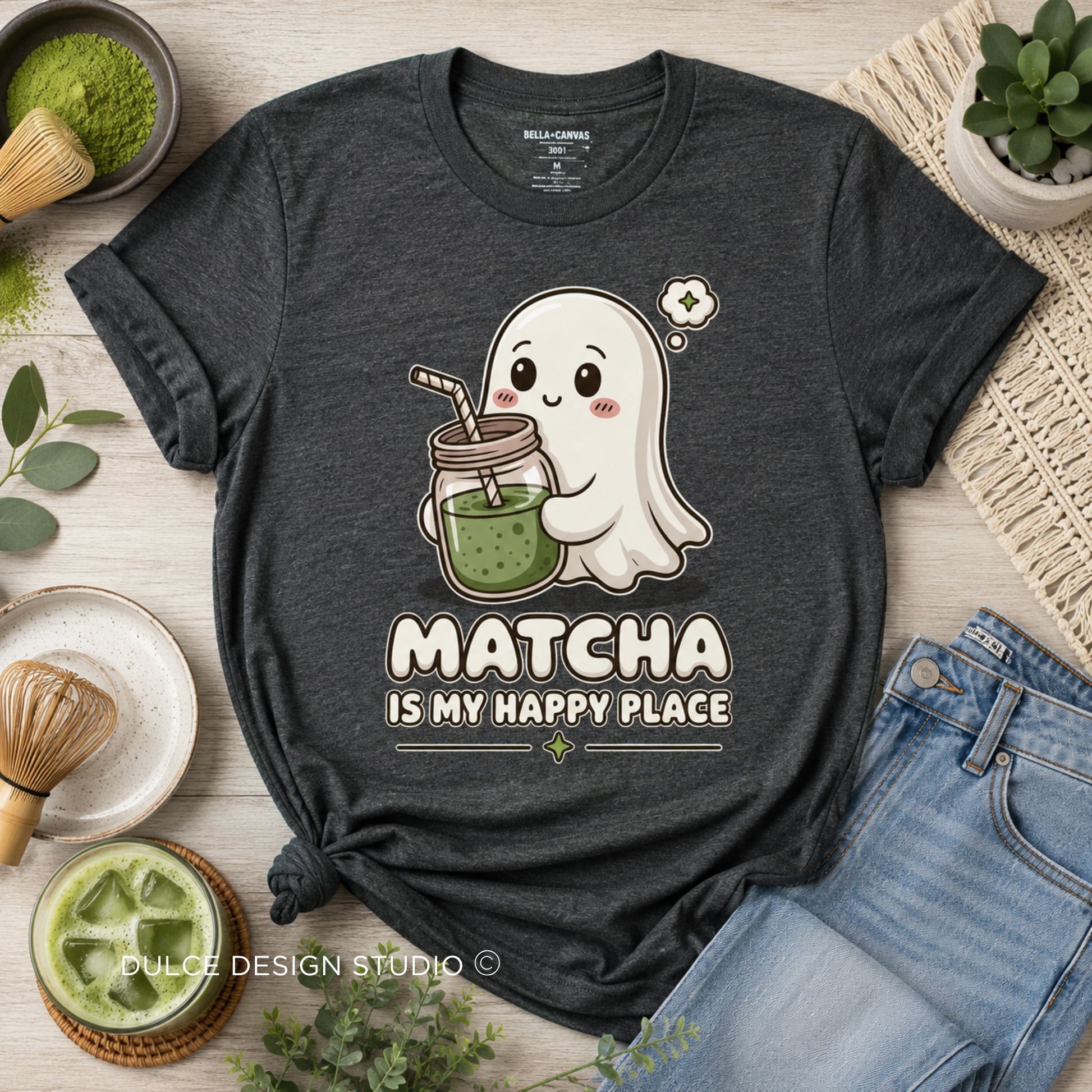 A dark heather gray graphic t-shirt laid flat on a light wood background, surrounded by matcha-themed props like a whisk, bowl, jeans, and a small plant. The shirt features a cute cartoon ghost holding a matcha drink with the text 'MATCHA IS MY HAPPY PLACE'.