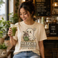 A woman in a cafe setting wearing a cream-colored graphic t-shirt with a cute ghost holding a matcha drink and the text 'MATCHA IS MY HAPPY PLACE'. She is holding a green iced matcha latte.