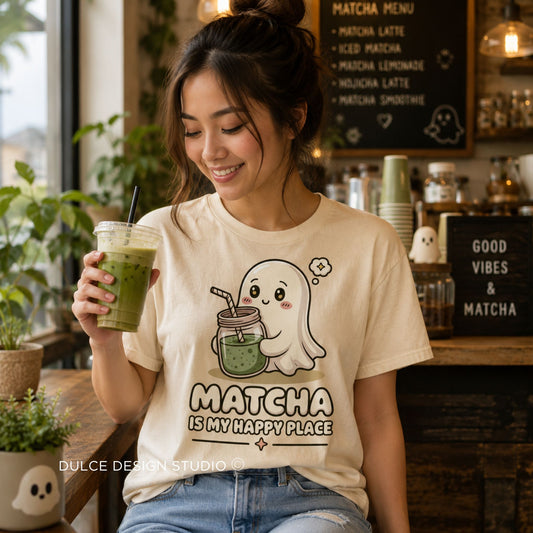 A woman in a cafe setting wearing a cream-colored graphic t-shirt with a cute ghost holding a matcha drink and the text 'MATCHA IS MY HAPPY PLACE'. She is holding a green iced matcha latte.