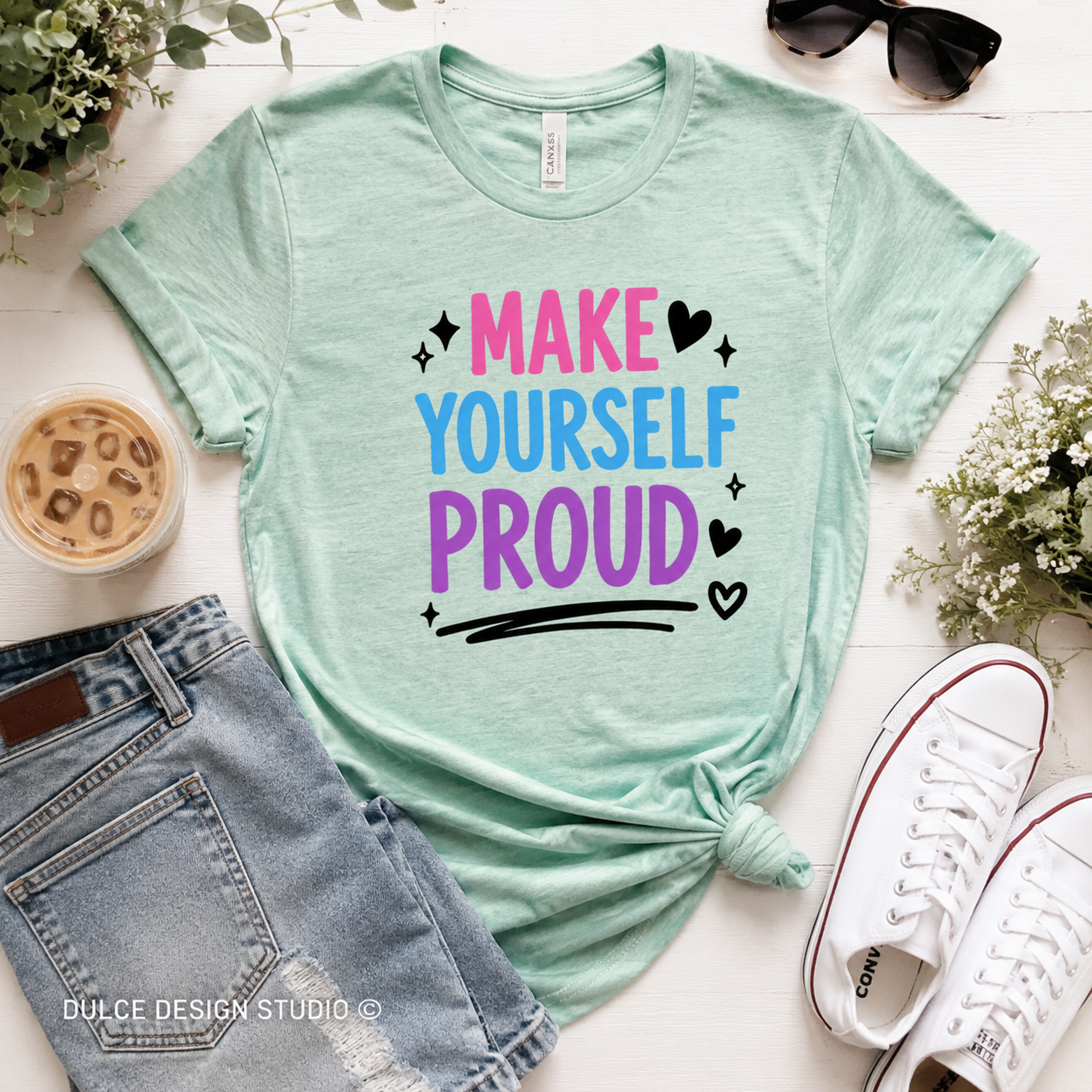 Mint green t-shirt with 'Make Yourself Proud' text, paired with jeans and sneakers on a light surface.