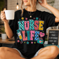 Woman wearing a black Nurse Life graphic tee with colorful nursing-themed design, holding a coffee mug