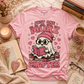 A pink graphic T-shirt laid flat on a cozy styled surface with books, a candle, a mug, glasses, and dried flowers. The shirt features a cartoon ghost wearing a pink beanie and reading a book, with the text 'JUST ONE MORE CHAPTER' and small hearts and sparkle accents.