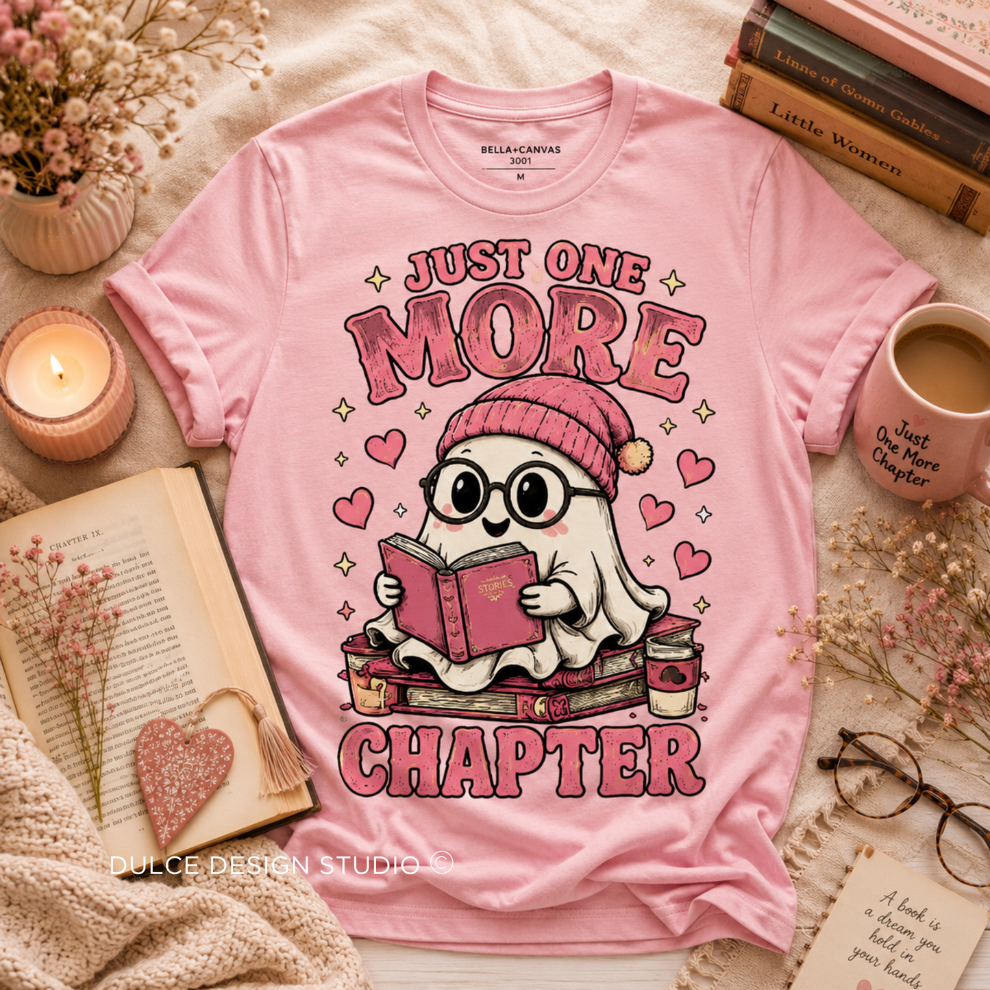 A pink graphic T-shirt laid flat on a cozy styled surface with books, a candle, a mug, glasses, and dried flowers. The shirt features a cartoon ghost wearing a pink beanie and reading a book, with the text 'JUST ONE MORE CHAPTER' and small hearts and sparkle accents.