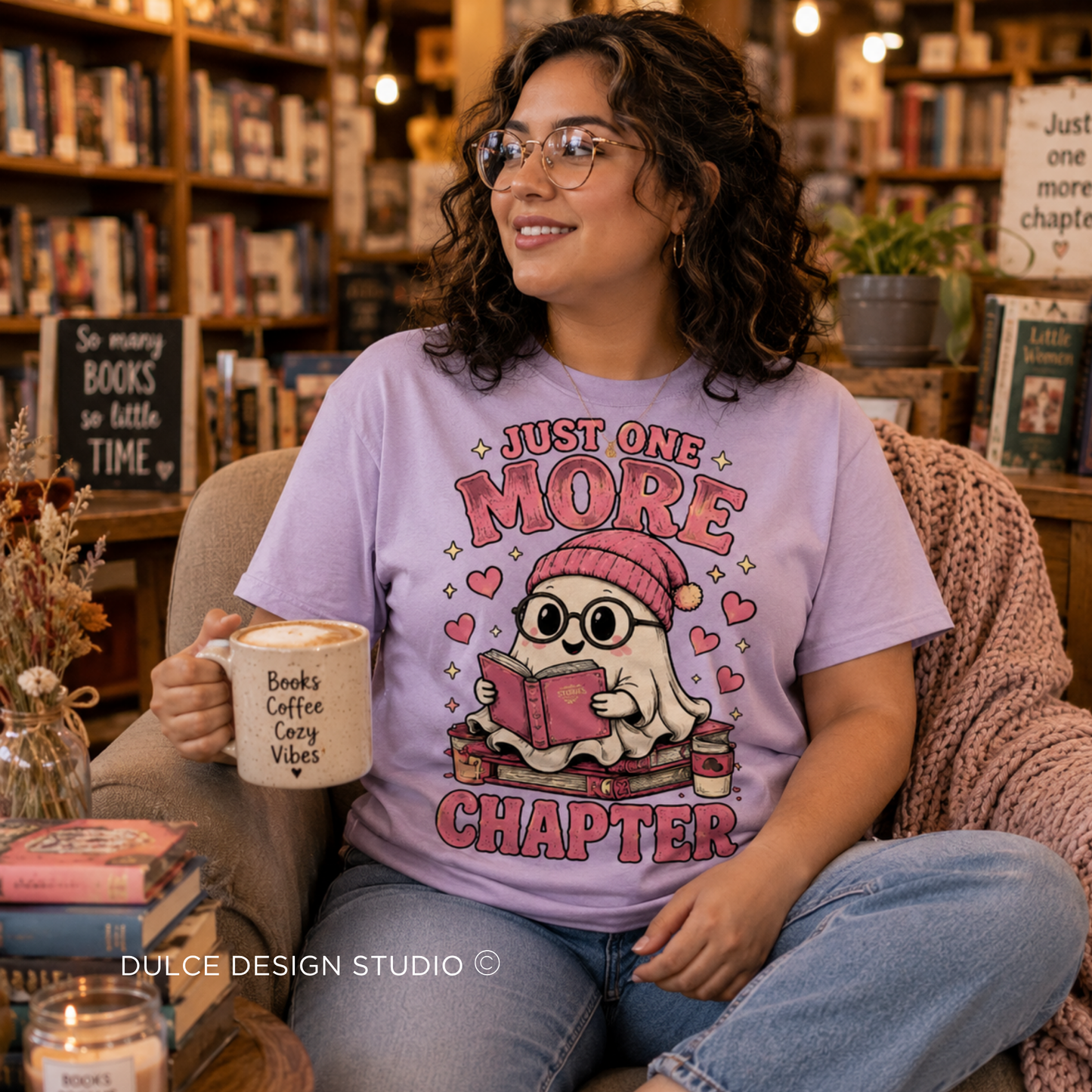 A woman wearing glasses sits in a cozy bookshop armchair, wearing a lavender graphic t-shirt that reads 'JUST ONE MORE CHAPTER' with a cute ghost character reading a book. She holds a mug that says 'Books Coffee Crazy Vibes'.
