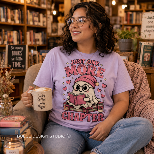 A woman wearing glasses sits in a cozy bookshop armchair, wearing a lavender graphic t-shirt that reads 'JUST ONE MORE CHAPTER' with a cute ghost character reading a book. She holds a mug that says 'Books Coffee Crazy Vibes'.