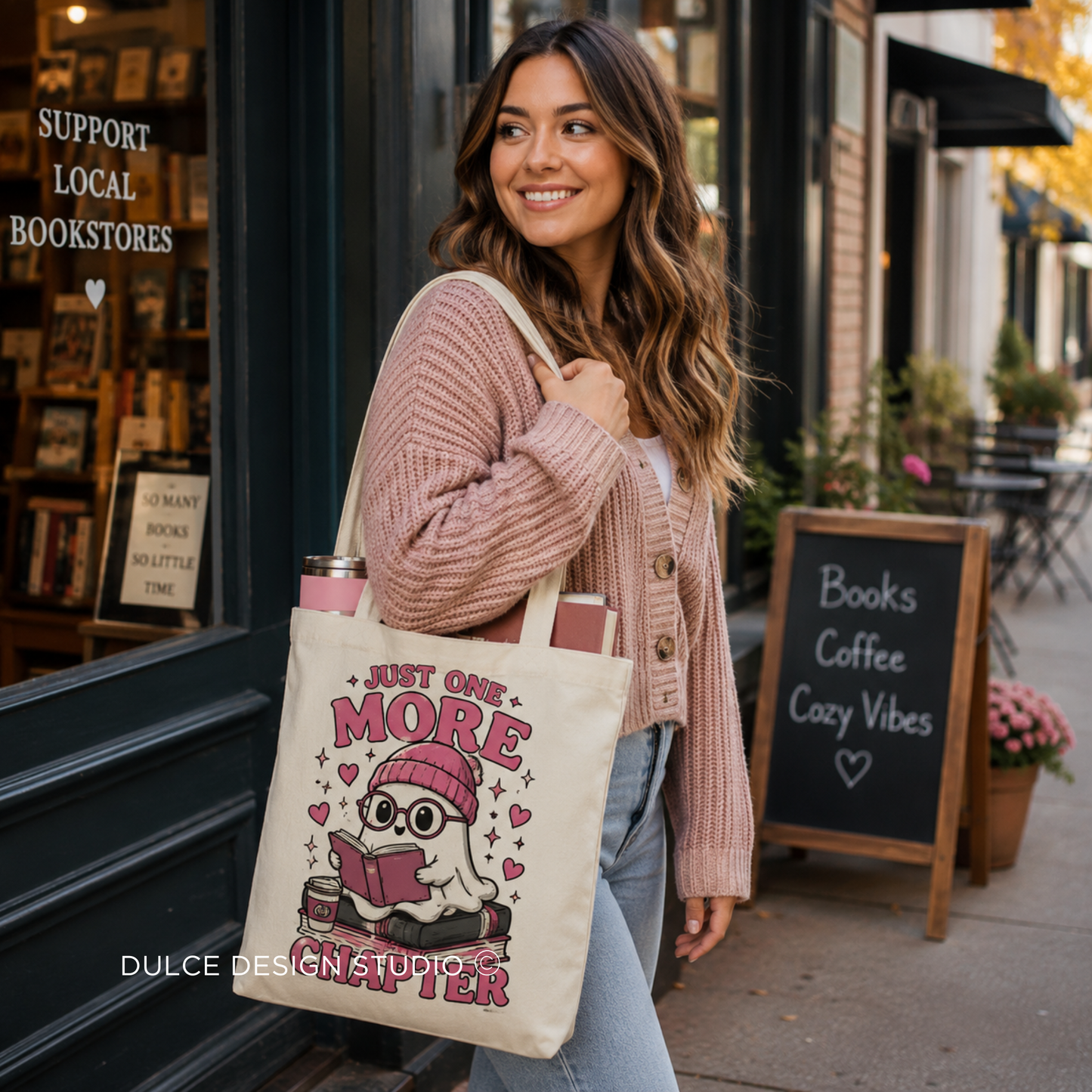 A woman in a pink cardigan and jeans stands on a city sidewalk carrying a cream canvas tote bag with a cartoon ghost reading a book and the text 'JUST ONE MORE CHAPTER'. The background shows a bookstore storefront and a chalkboard sign reading 'Books Coffee Cozy Vibes'.