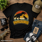 Black t-shirt with 'Papasaurus' design, sunglasses, mug, jeans, and sneakers on a wooden surface.
