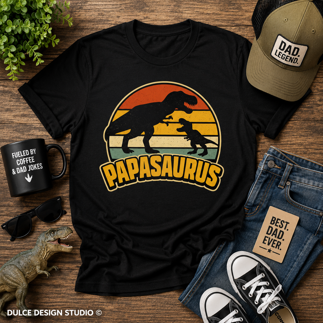 Black t-shirt with 'Papasaurus' design, sunglasses, mug, jeans, and sneakers on a wooden surface.