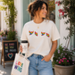 Woman wearing a white t-shirt with colorful designs, holding a coffee cup and a tote bag with motivational text.