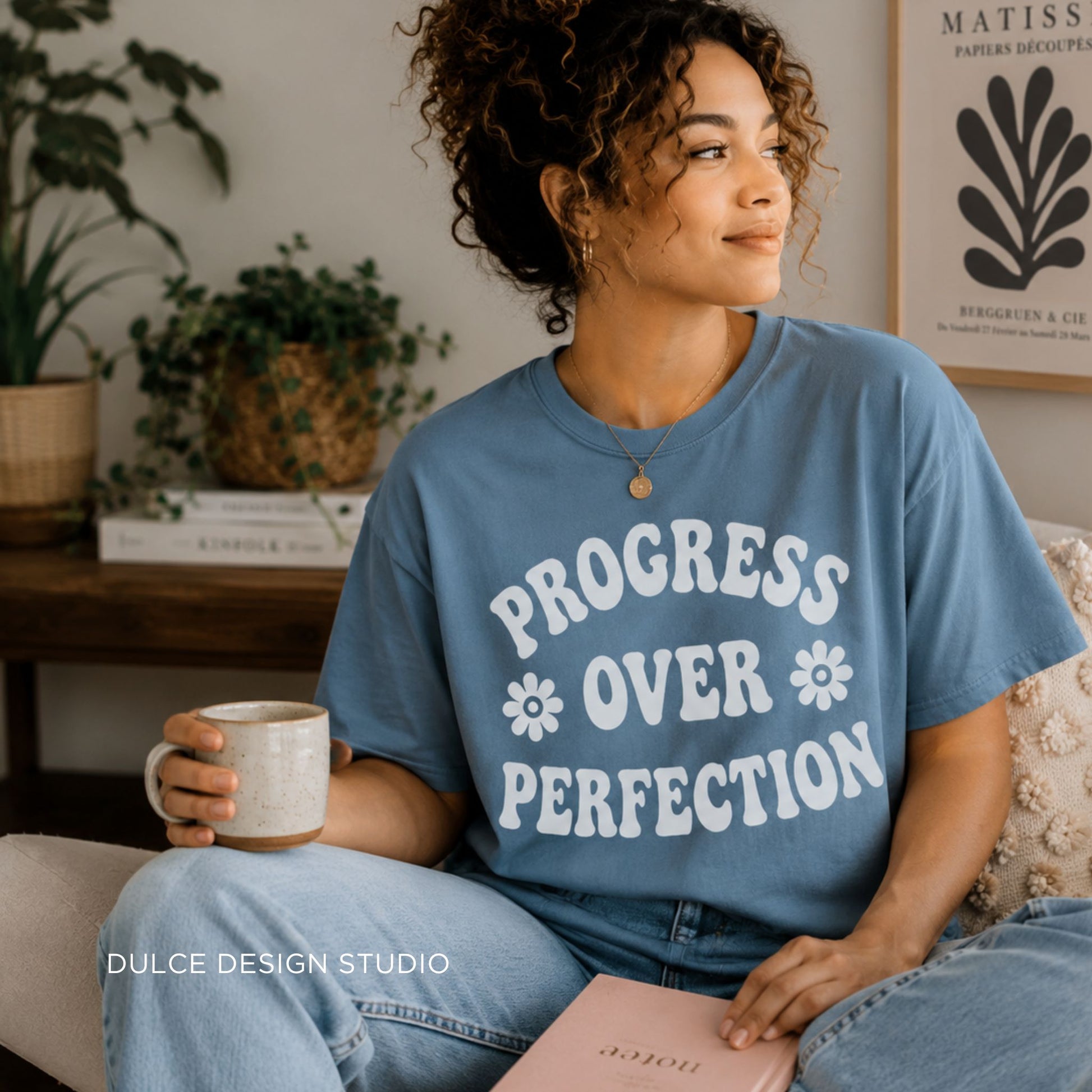 Person wearing a blue t-shirt with 'Progress Over Perfection' text, holding a mug and a book in a cozy room.