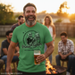 Man wearing a green t-shirt with a graphic design and text, holding a beer, standing outdoors with friends around a fire.