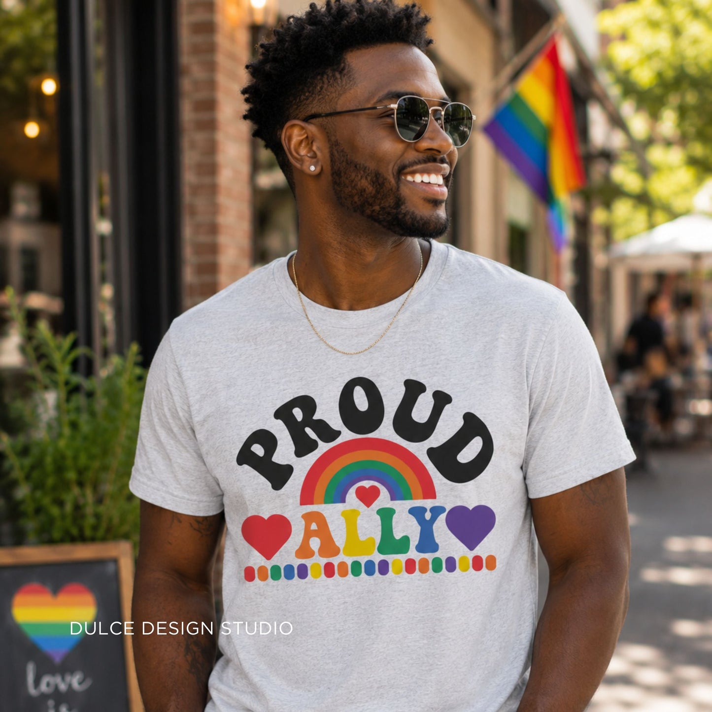 Man wearing a t-shirt with 'Proud Ally' design on a street with a rainbow flag in the background.