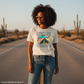 Woman wearing a t-shirt with a graphic design and text, standing on a desert road.