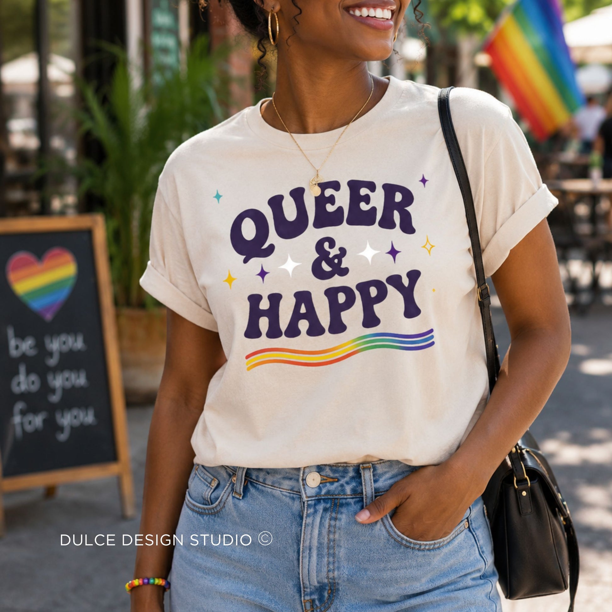 Person wearing a 'QUEER & HAPPY' t-shirt with rainbow elements in an outdoor setting.