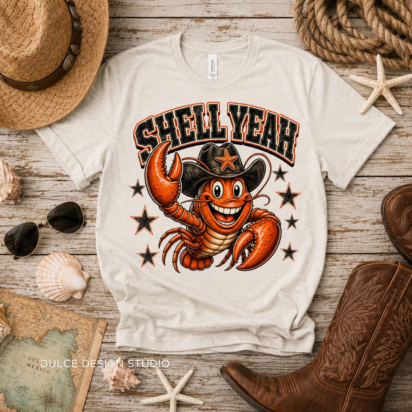 White t-shirt with a lobster wearing a cowboy hat and 'Shell Year' text, surrounded by coastal items on a wooden surface.