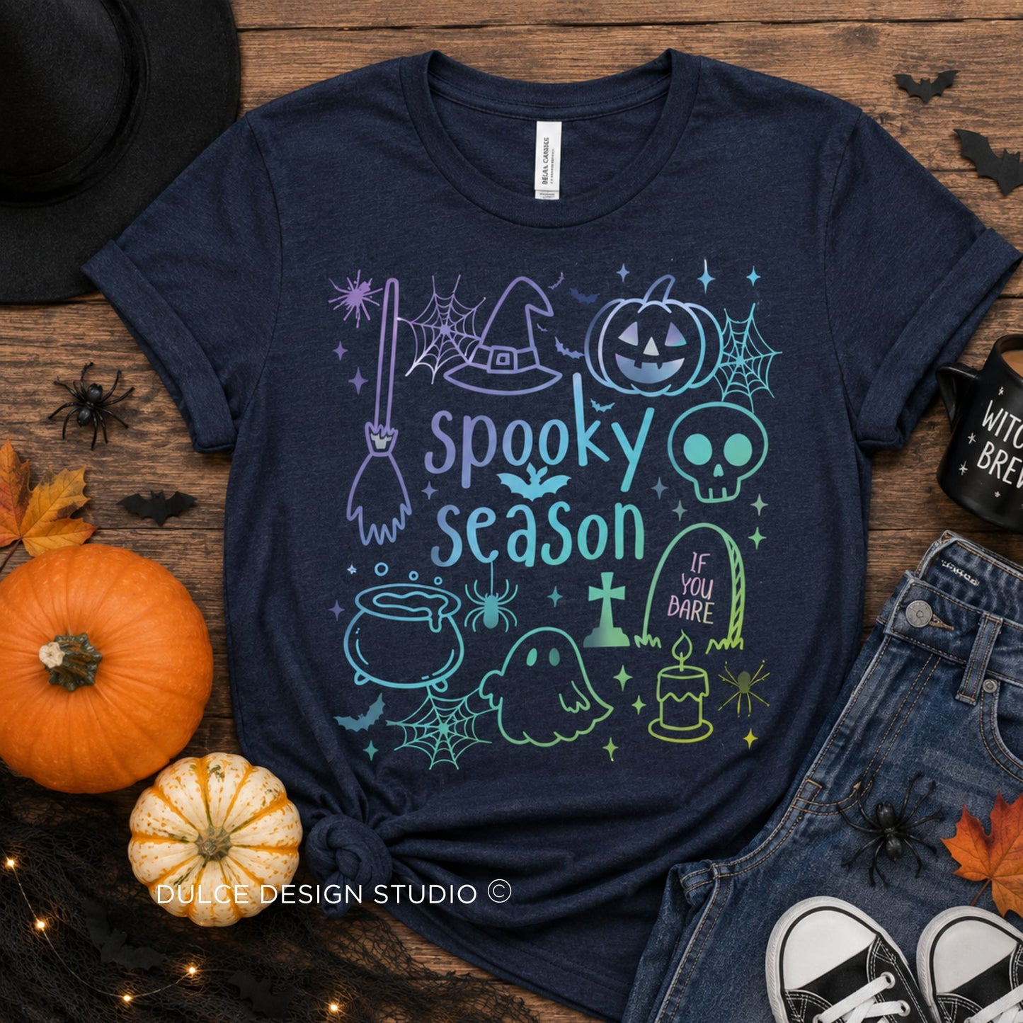 A navy blue graphic t-shirt featuring a 'Spooky Season' Halloween design with a witch hat, pumpkin, skull, cauldron, ghost, and tombstone in light blue and mint green. The shirt is styled in a flat-lay arrangement on a rustic wooden background with a black hat, orange pumpkins, denim shorts, and a black coffee mug.
