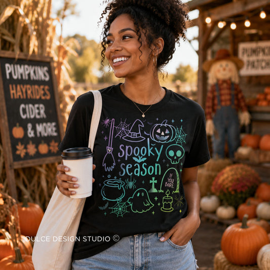 A woman standing in a fall-themed outdoor shop holding a coffee cup, wearing a black graphic t-shirt with a colorful 'Spooky Season' design featuring pumpkins, ghosts, and a witch hat.