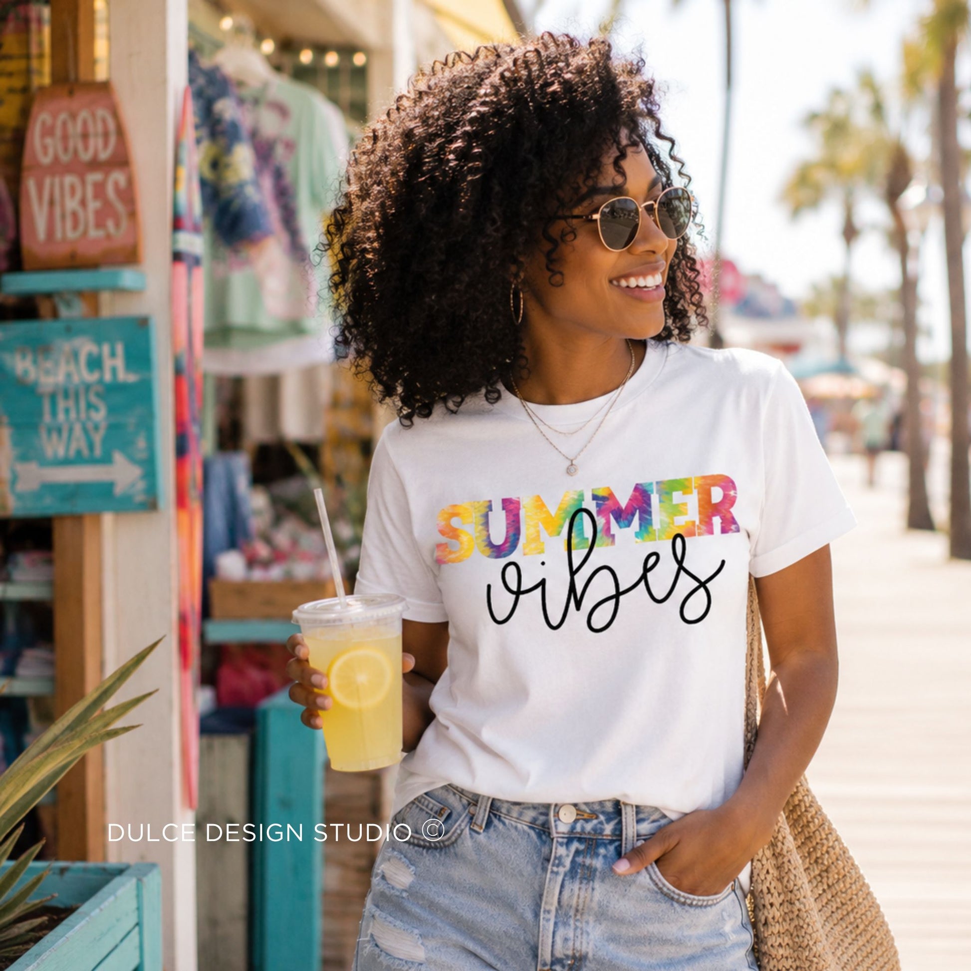 Woman wearing a 'Summer vibes' t-shirt holding a drink, with palm trees and a beach store in the background.