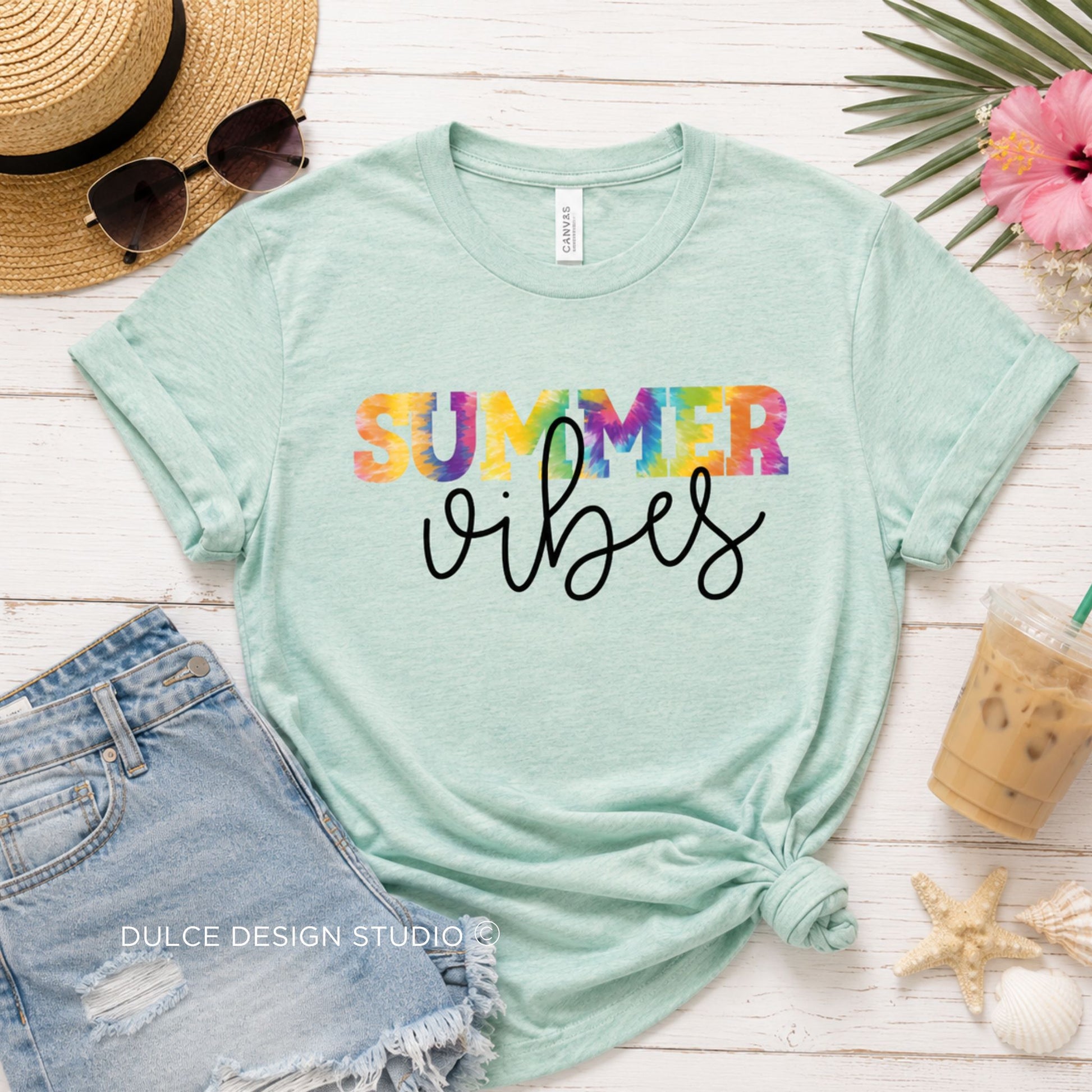 Mint green t-shirt with 'Summer vibes' text on a wooden surface with sunglasses, shorts, and a drink.