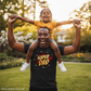Man wearing a 'Super Dad' shirt holding a child on his shoulders in a park.