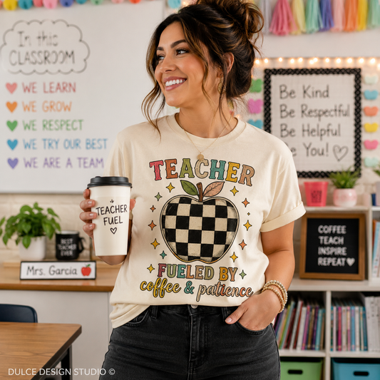 A woman in a classroom setting wearing a cream-colored graphic t-shirt that reads 'TEACHER FUELED BY coffee & patience' with a checkerboard apple design. She is holding a matching coffee cup and smiling.