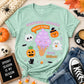 A mint green graphic t-shirt styled in a flat-lay arrangement with Halloween props, including a pumpkin, skull, black cat figurine, jeans, and a coffee cup. The t-shirt features a cute illustration of a purple ice cream cone with a witch hat pumpkin on top, surrounded by skulls, ghosts, bats, and candy corn, with the text 'HAUNTINGLY ADORABLE TOO CUTE TO FEAR! BOOS! BOOTS!'.