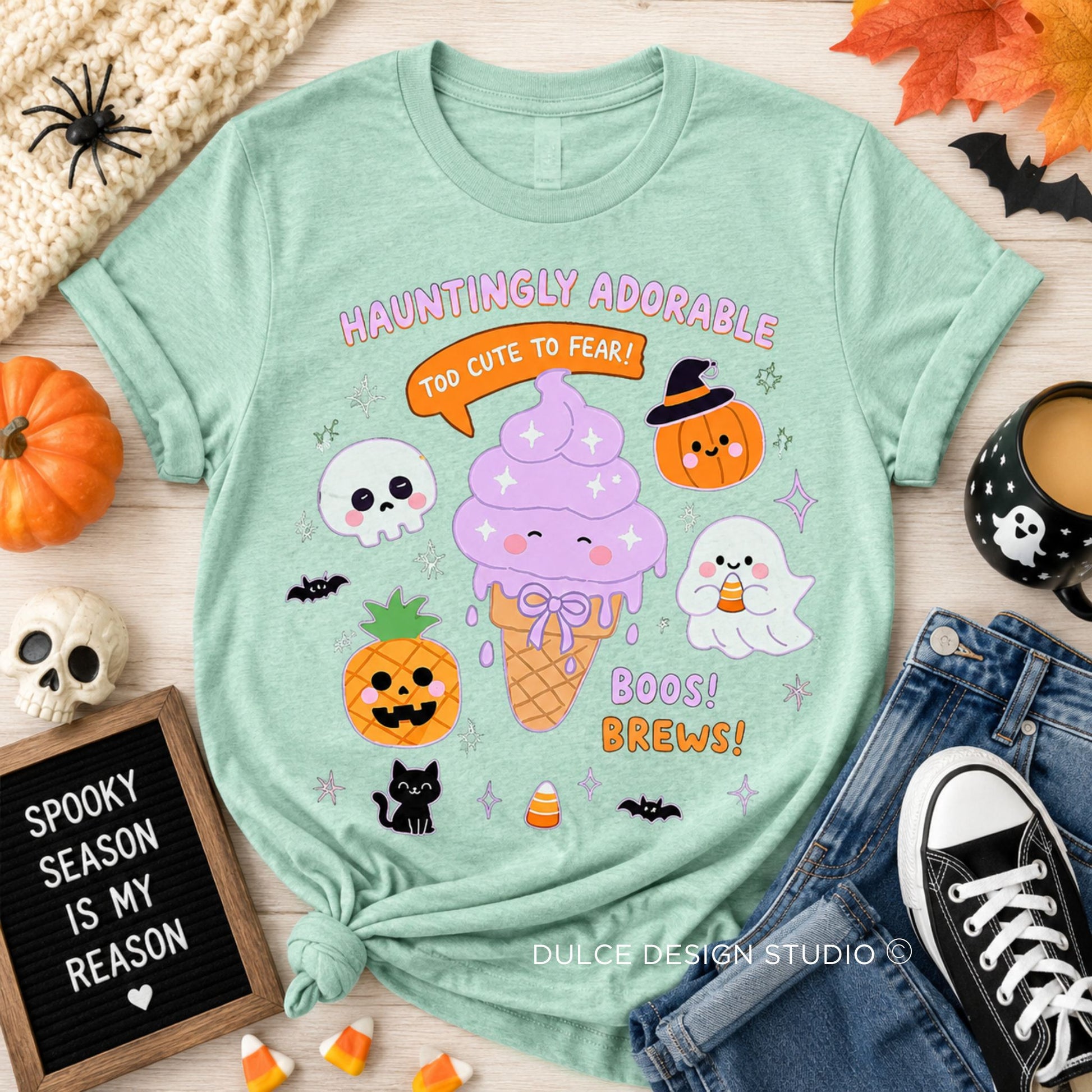 A mint green graphic t-shirt styled in a flat-lay arrangement with Halloween props, including a pumpkin, skull, black cat figurine, jeans, and a coffee cup. The t-shirt features a cute illustration of a purple ice cream cone with a witch hat pumpkin on top, surrounded by skulls, ghosts, bats, and candy corn, with the text 'HAUNTINGLY ADORABLE TOO CUTE TO FEAR! BOOS! BOOTS!'.